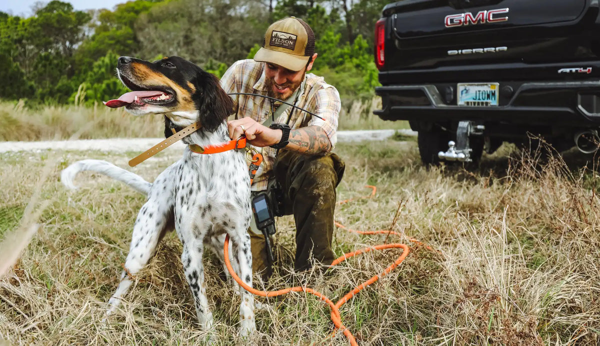 A man holding his bird dog kneeling in grass with a pickup in the background.