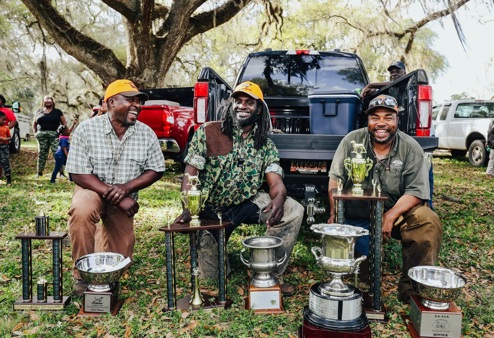 Three men posing with their trophies with several pickup trucks in the background.