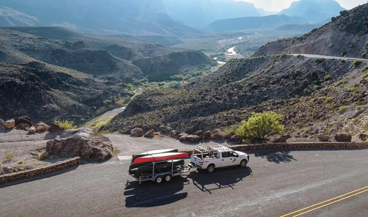 A pickup hauling a trailer with canoes down on a mountain road with a scenic landscape in the background