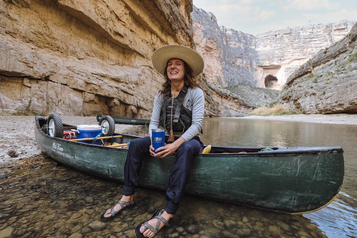 A woman sitting in a canoe holding a blue B&W cup, surrounded by rocky canyon walls.