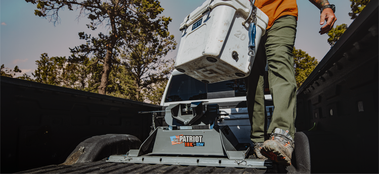 A man standing in the bed of a pickup truck loads a white cooler alongside a Patriot rail-mounted fifth wheel hitch, with trees and a blue sky in the background.