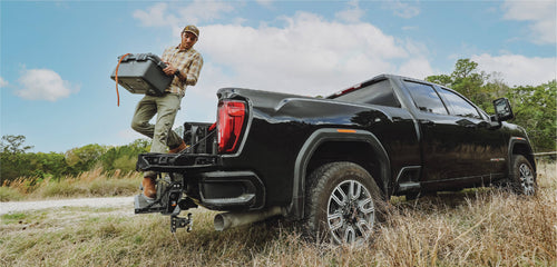 A man stepping off of a pickup bed onto a multipro tailgate and the pickup is parked in tall grass.