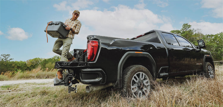 A man stepping off of a pickup bed onto a multipro tailgate and the pickup is parked in tall grass.