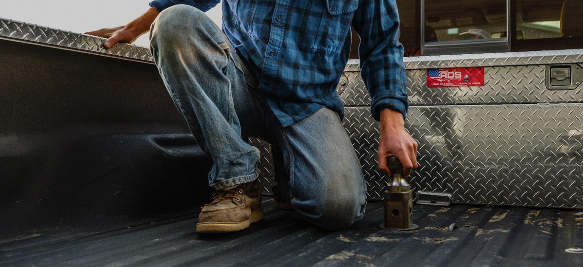 A man kneeling in a truck bed with a toolbox, wearing a blue plaid shirt and jeans inserting a gooseneck ball into a turnoverball hitch