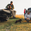 A man sits in the bed of a pickup truck holding a brown puppy while another man kneels on the grass nearby, with a second puppy running between them in an outdoor field at sunset.