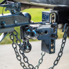 Close-up of a chrome tow & stow adjustable trailer hitch with safety chains attached to a vehicle frame. The hitch features a blue 'BW' logo, with a blurred outdoor background of green grass and gray pavement.
