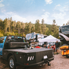 A black flatbed truck with a companion flatbed fifth wheel hitch, shown at an outdoor camping site with RVs, tents, trees, and people in the background.