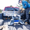 A man in a blue hoodie and black cap loads a B&W companion puck-mount fifth wheel hitch into the bed of a red pickup truck outdoors on a sunny day.