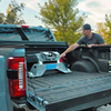 A man in a black cap and gray t-shirt reaches in the bed of a light blue pickup truck with a Companion puck-mounted fifth wheel hitch installed. The truck is parked outdoors with trees and a blue sky in the background.