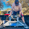 A man in a light-colored long-sleeve shirt, black shorts, and a dark cap is loading a B&W companion puck-mounted fifth wheel hitch into the bed of a pickup truck outdoors under a blue sky with trees in the background.