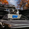 A B&W companion single-point fifth wheel hitch, mounted on the bed of a black pickup truck in an outdoor setting with autumn trees in the background.