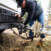 A man in a black cap and dark jacket is attaching a heavy-duty dual-ball pintle tow & stow trailer hitch with safety chains to a white trailer outdoors on grassy ground.