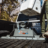 A man standing in the bed of a pickup truck loads a white cooler alongside a Patriot rail-mounted fifth wheel hitch, with trees and a blue sky in the background.