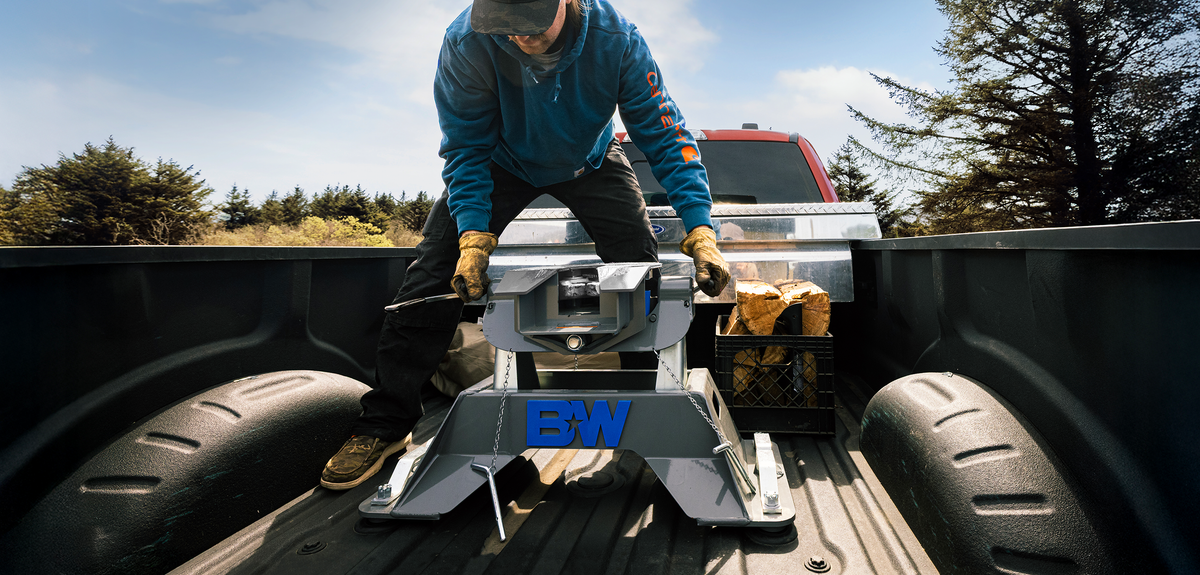 A man in a blue jacket and black pants is installing a companion oem fifth wheel hitch into the bed of a pickup truck outdoors. The trailer has a blue 'B&W' logo on the front.