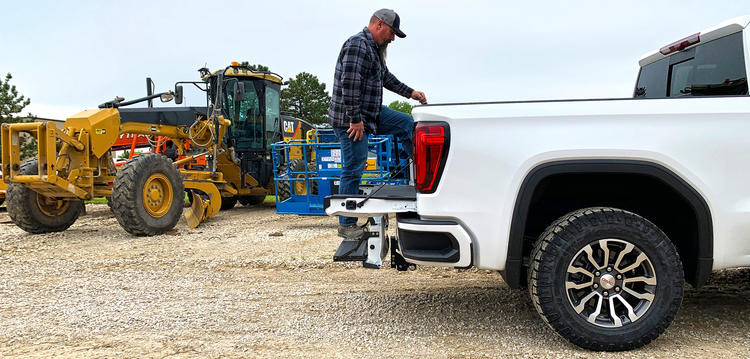 A man in a plaid shirt and cap steps up on a  multipro tailgate on a white pickup truck, while the b&w multipro-multiflex hitch clears the tailgate. This is a construction setting with heavy machinery in the background.