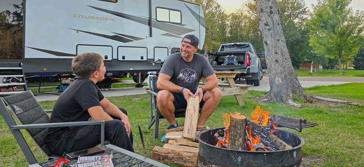 a dad and his son sit beside a campfire and there's an rv and a pickup truck with a companion fifth wheel hitch in the bed.
