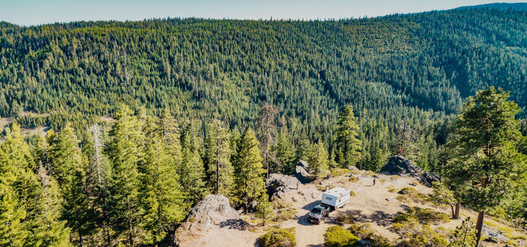 a pickup hauling a travel trailer that's parked on a mountain ledge with a landscape of tall pine trees