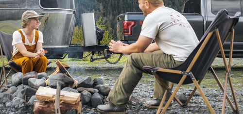 Two people sitting by a campfire with a camper, b&w weight distribution hitch, and truck in the background