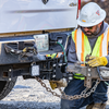 A construction worker wearing a high-visibility safety vest, yellow gloves, and a hard hat attaches a trailer to a B&W heavy-duty receiver hitch hitch in an outdoor setting.