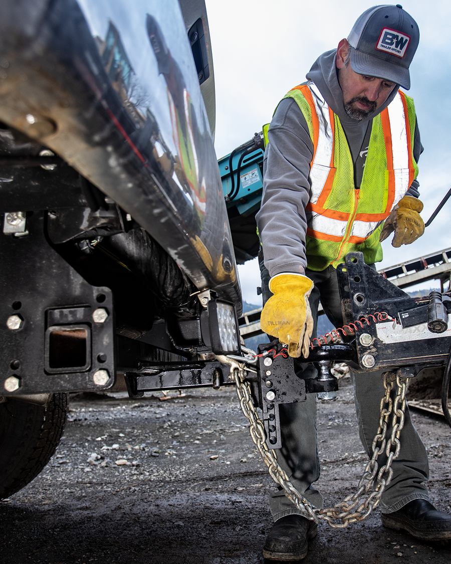 A construction worker wearing a high-visibility safety vest, yellow gloves, and a cap with a 'B&W' logo attaches a trailer to a heavy-duty receiver hitch hitch in an outdoor industrial setting.