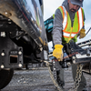 A construction worker wearing a high-visibility safety vest, yellow gloves, and a cap with a 'B&W' logo attaches a trailer to a heavy-duty receiver hitch hitch in an outdoor industrial setting.