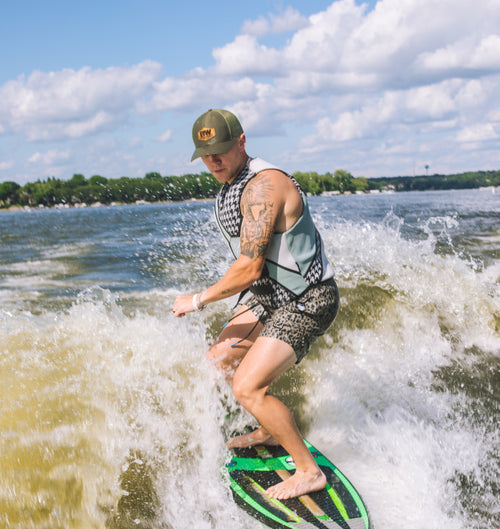 a man wearing a lifejacket and a B&W leather patch hat while riding a surfboard in the wake of a boat. 