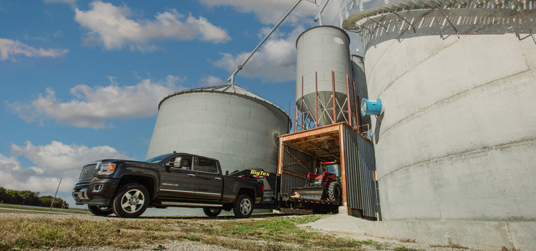 a pickup truck backing up a gooseneck trailer with a tractor and front end loader backed into a shed surrounded by grain bins