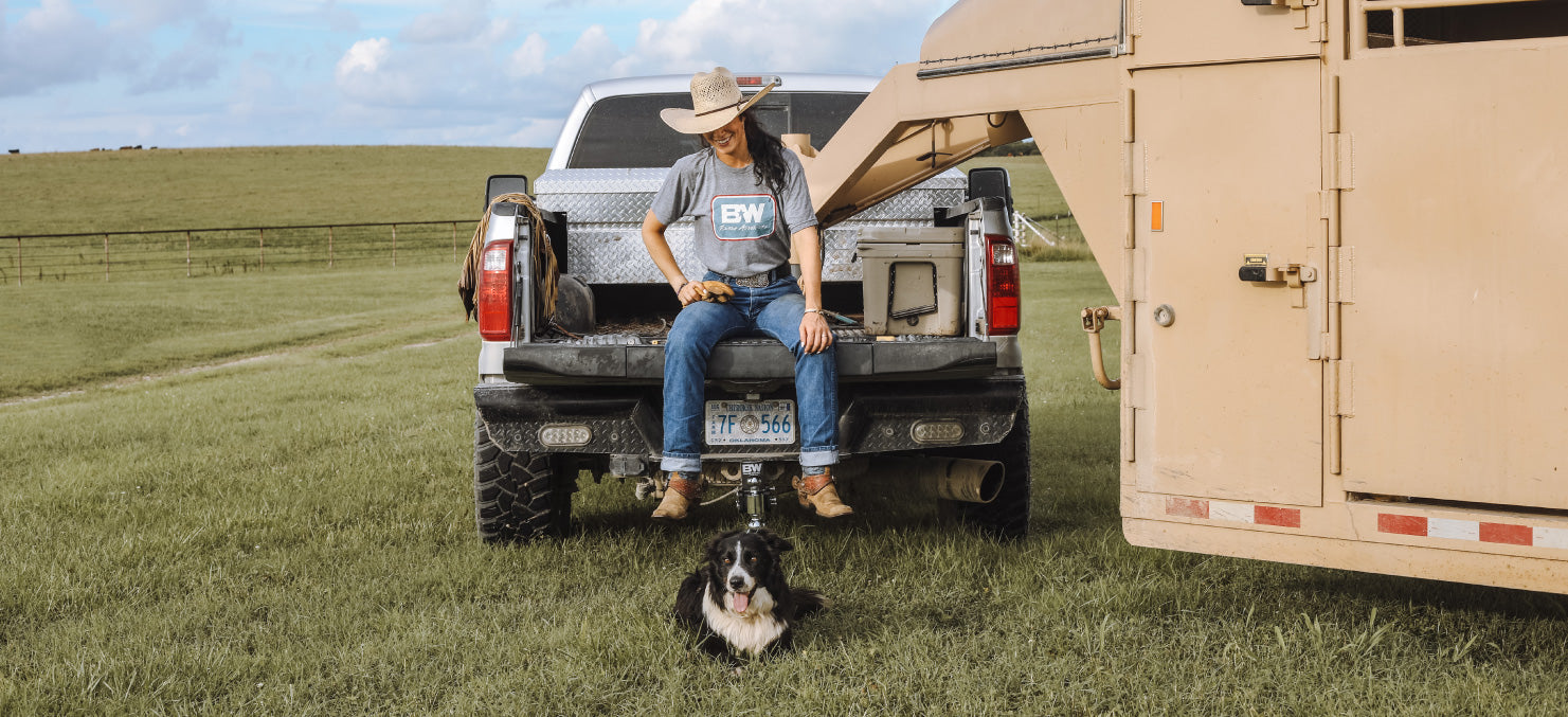 a woman sitting on a tailgate of a pickup with a dog laying in the grass below her. There is a gooseneck trailer attached to the pickup and a Tow & Stow hitch on the truck.