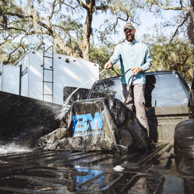 Man standing in back of truck spraying a hose and washing out the truck bed with fifth wheel hitch and an RV fifth wheel camper in background.